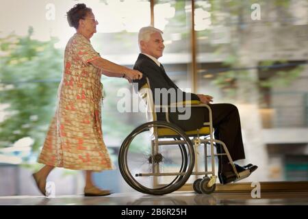 Blick auf eine ältere Frau, die einen Mann auf den Rollstuhl drückt. Stockfoto