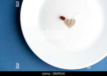 Granulate aus einer Kapsel auf eine weiße Platte in Form eines Herzens auf einem klassischen blauen Hintergrund gegossen. Das Herzsymbol besteht aus Granulat Stockfoto