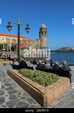 Collioure, Frankreich; Menschen sitzen genießen warmen Frühlingstag in Mittelmeer-Küstendorf mit Kirche, Notre-Dame-des-Anges im Hintergrund Stockfoto