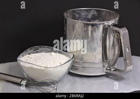 Eine Schüssel mit Mehl neben einem mechanischen Sieb und einem Schneebesen auf Backpapier und dunklem Hintergrund Stockfoto