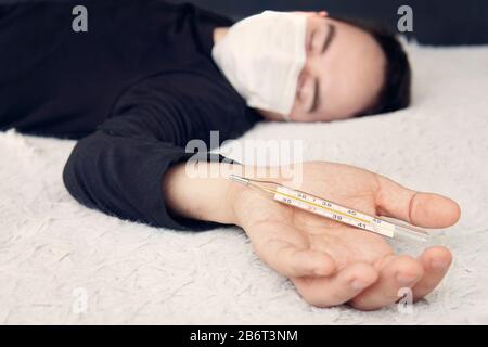 Der Kranke, der eine schützende medizinische Maske im Bett trägt, liegt auf dem Bett und hält ein Thermometer mit hoher Temperatur in der Hand. Thermometer Stockfoto