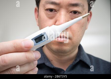 Ein asiatischer Mann mittleren Alters, der das Thermometer ängstlich in der Hand betrachtet. Stockfoto