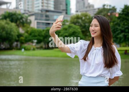 Fröhliches junges, wunderschönes asiatisches Teenager-Mädchen, das selfie im Park mitnimmt Stockfoto