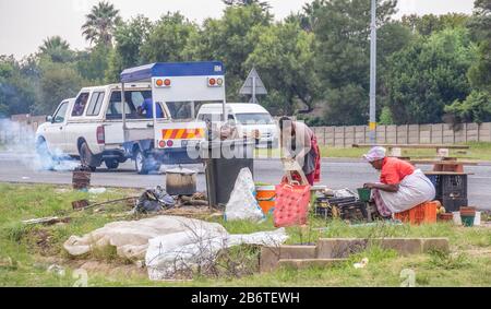 Alberton, Südafrika - unidentifizierte schwarze Afrikanerinnen verkaufen geröstete Mealies und frische Mangos aus einem provisorischen Straßenmeisterei Stockfoto