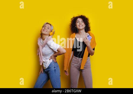 Fröhliche kaukasische Schwestern mit hübschen lockigen Haaren hören Musik mit Kopfhörern, während sie auf einem gelben Hintergrund posieren Stockfoto