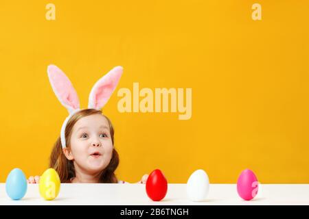 Osterkonzept. Entzückt überraschte glückliches Kind in den Ohren eines Hünenbrunnens auf gelbem Hintergrund. Kleines Mädchen sucht vom Tisch auf dem Copy-Platz nach Stockfoto