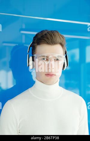 Porträt eines ernsthaften jungen Mannes mit moderner technischer Brille und drahtlosen Kopfhörern, die gegen die blaue Wand stehen Stockfoto