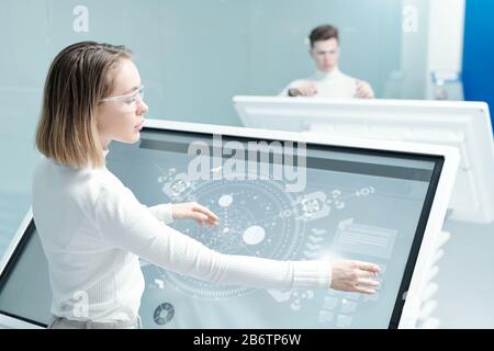 Junge Frau in Brille arbeitet mit digitalem Schema auf interaktivem Display im modernen Büro Stockfoto