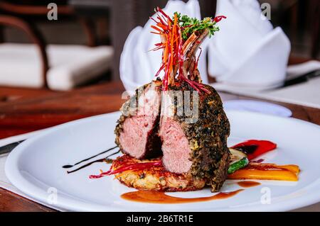 Gebratenes Lamb-Schopfsteak mit knuspriger Kräuterkruste und gegrilltem Gemüse auf weißem Teller am Esstisch Stockfoto