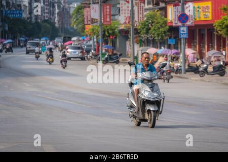 Yangshuo, China - August 2019: Älterer Mann, der auf einem Roller auf einer breiten Straße in der Yangshuo-Stadt in der Provinz Guangxi fährt Stockfoto