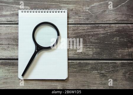 Notepad mit einem leeren weißen Blatt in einem Schachpapier liegt mit Lupe auf dem Hintergrund von Holzbrettern. Stockfoto