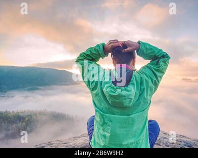 Junge Frau sitzt auf einem Berg den Tag feiern. Junge Dame Wanderer sitzen in blau grün Windjacke mit roten Rucksack auf scharfe Klippe Stockfoto