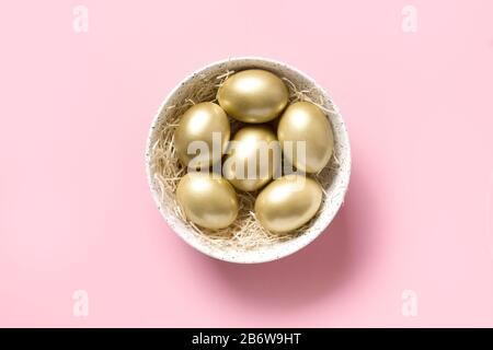 Ostergoldenes Hühnerei in weißer Schüssel auf pinkfarbenem Hintergrund. Ansicht von oben. Minimales Konzept. Stockfoto