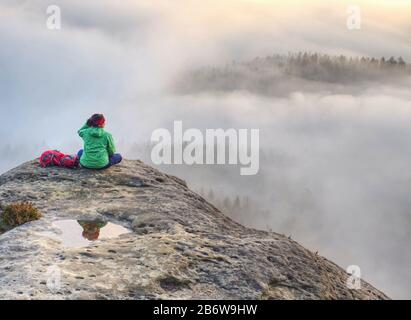 Grüne Jacke Frau sitzt penibel am Rand eines Felsens und betrachtet die nebligen Wolken. Stockfoto