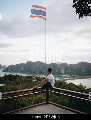 Mann, der auf einem Felsvorsprung steht und auf der schönen thailändischen Insel mit der thailändischen Flagge winkt Stockfoto