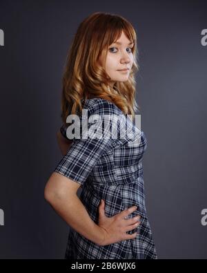 Ein Mädchen mit goldenen Locken sieht eine halbe Umdrehung aus. Sie trägt ein blau kariertes Kleid. Die Hand liegt auf der Hüfte. Studio gedreht. Stockfoto