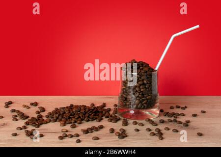 Whiskyglas voller gerösteter Kaffeebohnen mit Strohhalm, das auf einem Holzbrett vor rotem Hintergrund steht Stockfoto