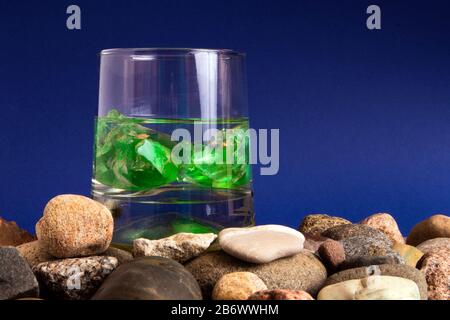 Glas mit Wasser und zwei grünen Edelsteinen im Inneren, die auf einem Kieselhaufen stehen Stockfoto