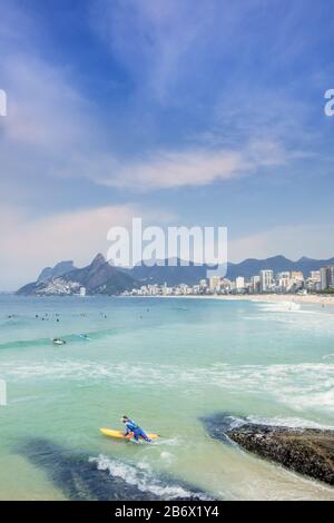 Ein männlicher Surfer, der am Arpoador-Felsen mit Ipanema-Strand, Nachbarschaft und den Dois Irmaos-Bergen im Hintergrund in den Atlantik eindringt Stockfoto