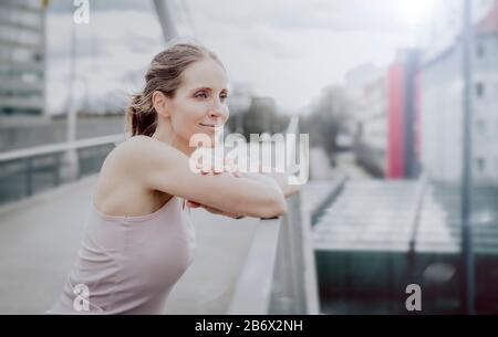 Fröhliche junge Frau ruht nach dem Joggen in der Stadt und lehnt sich an ein Geländer. Die Glückliche junge Frau ruht nach dem Joggen in der Stadt und stützt sich auf Stockfoto