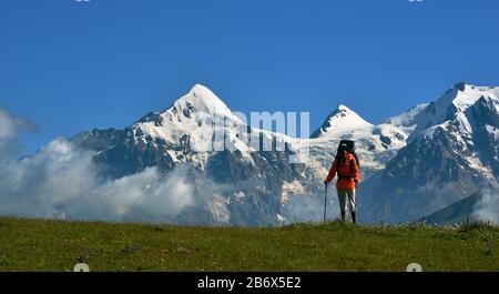 Tourist mit hellorangefarbenem Windschutzschalter mit Stöcken gegen schneebedeckten Oberseiten. Alpine Wiese steht im Vordergrund. Kaukasusgebirge (Tviber pe Stockfoto