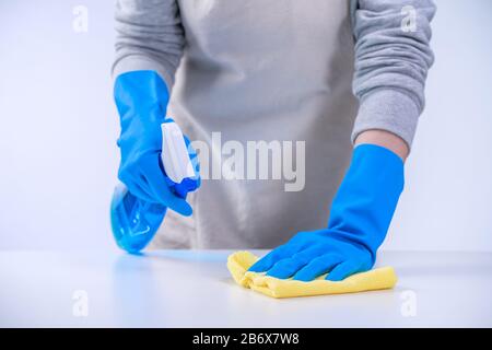Die Haushälterin der jungen Frau putzt den weißen Tisch in der Schürze mit blauen Handschuhen, Spritzreiniger, nassem gelbem Lappen, Nahaufnahme, Kopierraum, leerer Designkon Stockfoto