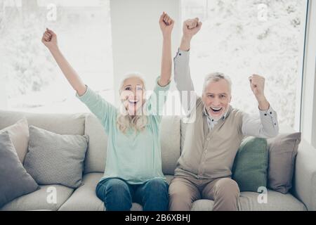 Portrait heiterer verheirateter Menschen in tealbraunen Pullovern, die Fäuste aufziehen, die schreien, ja sitzende Divan-Wohnung Stockfoto