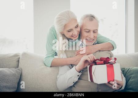 Porträt der niedlichen Großeltern gekleideten Pullover, die das Paket öffnen und die Sitzgelegenheit aussehenden Divan Zimmer-Wohnung halten Stockfoto