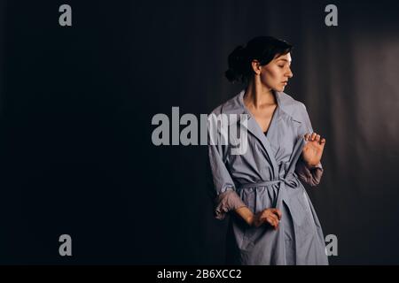 Mädchen mit Pferdeschwanz straffen den Gürtel eines blauen Grabenmantels. Studio mit leerem Raum aufgenommen. Mädchen mit Pferdeschwanz straffen den Gürtel eines blauen Tes Stockfoto