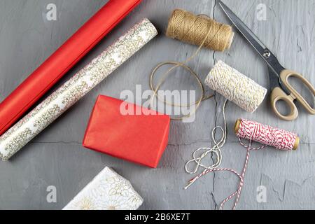 Vorbereitung auf die Verpackung von Geschenken, auf dem Tisch aufgelegtes rot-weißes Verpackungspapier, Fäden verschiedener Farben für Wicklung und Schere Stockfoto
