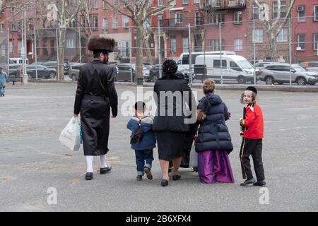 Eine Satmar-Familie geht mit ihren Kindern in Purim-Kostümen durch einen Schulhof. Abseits der Lee Ave. In Williamsburg, Brooklyn, New York. Stockfoto