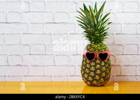 Eine Ananas, die ein Paar pinkfarbene herzförmige Sonnenbrillen auf gelber Oberfläche vor einem weißen Backstein-Wandhintergrund mit etwas Leerstand trägt Stockfoto