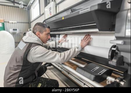 Der Bediener des Technikers wechselt die Papierrolle auf einem großen hochwertigen Industriedrucker und Plottergerät im digitalen Druckereibüro Stockfoto