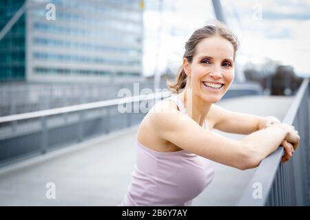 Fröhliche junge Frau ruht nach dem Joggen in der Stadt und lehnt sich an ein Geländer. Die Glückliche junge Frau ruht nach dem Joggen und stützt sich auf ein Geländer. Stockfoto