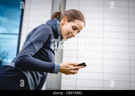 Junge, sportliche Frau macht dem Joggens eine Pause mit griffiger Hand. Junge, sportliche Frau macht sich eine Pause mit einem Handy in der Hand. Stockfoto