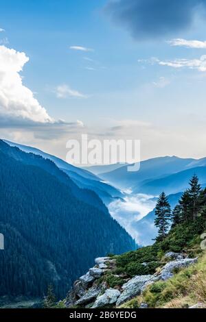 Bergfelsen und Bäume im Vordergrund mit nebelig bewaldeten Bergen im Hintergrund und puffigen Wolken vor dem blauen Himmel Stockfoto