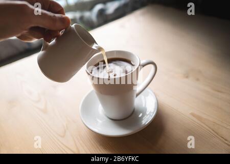 Milch, die aus einer kleinen Kanne in eine Tasse Kaffee oder Tee gegossen wird. Die Kanne, der Becher und die Untertasse sind weiß. Sie befinden sich auf einem Holztisch, das Tageslicht ist Stockfoto