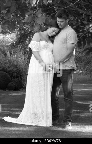 Schöne schwangere junge Frau mit ihrem Lebensgefährten, die mit ihren Armen nebeneinander steht, schwarz-weiß Stockfoto