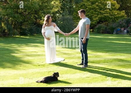 Schöne schwangere junge Frau, die ein langes weißes Kleid trägt und an einem schönen Sommertag Hand in Hand mit ihrem Partner geht Stockfoto