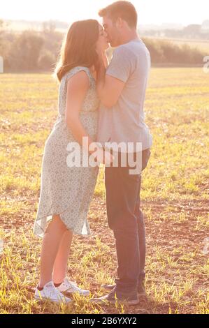 Schwangere junge Frau mit ihrem Partner küsst und kuschelt, steht an einem schönen Sommertag auf einem Feld Stockfoto