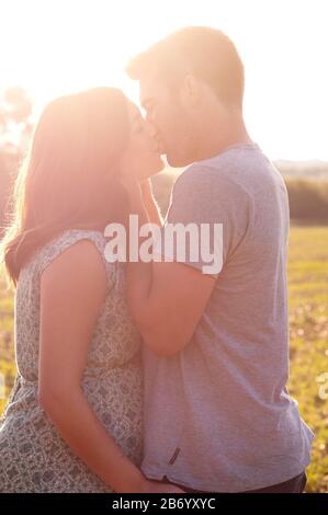 Schwangere junge Frau mit ihrem Partner küsst und kuschelt, steht an einem schönen Sommertag auf einem Feld Stockfoto