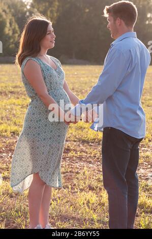 Schwangere junge Frau mit ihrem Partner, der die Hände hält, sich gegenseitig betrachtet und auf einem Feld steht Stockfoto