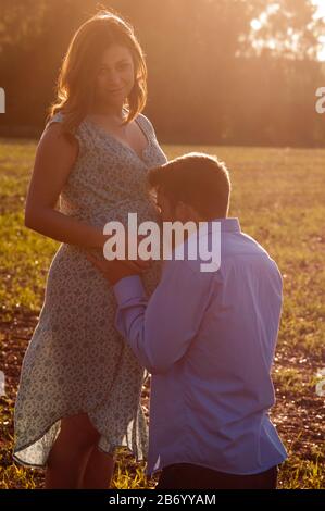 Schwangere junge Frau, die mit ihrem Partner auf einem Feld steht und ihren Bauch küsst Stockfoto