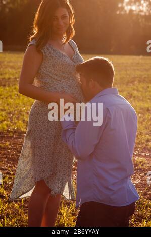 Schwangere junge Frau, die mit ihrem Partner auf einem Feld steht und ihren Bauch küsst Stockfoto