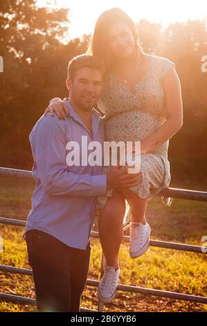 Schwangere junge Frau sitzt mit ihren Armen um ihren Partner auf einem Tor, hinter dem die Sonne untergeht Stockfoto