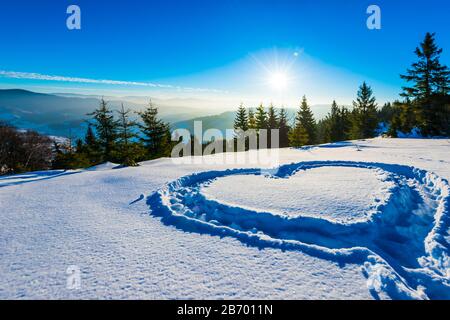 Herz trampelte auf dem Schnee mit Füßen in einer Schneeverwehung auf einem Hang mit herrlichem Blick auf den Nadelwald und die Bergketten auf einem sonnigen Frost Stockfoto