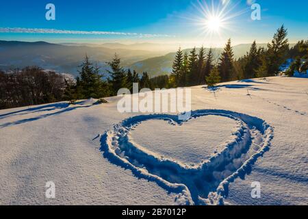 Herz trampelte auf dem Schnee mit Füßen in einer Schneeverwehung auf einem Hang mit herrlichem Blick auf den Nadelwald und die Bergketten auf einem sonnigen Frost Stockfoto