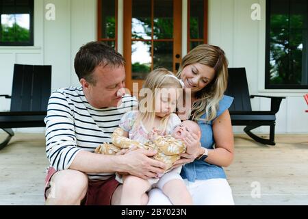 Ein junges Mädchen hält ihre neugeborene Schwester mit ihren Eltern auf einer Veranda Stockfoto