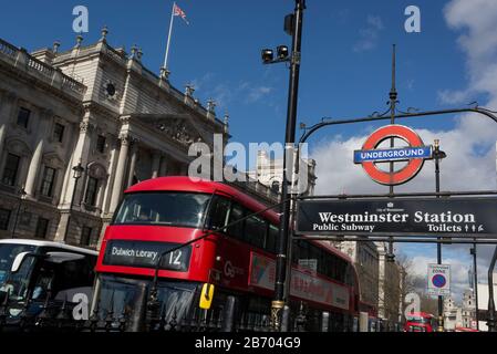 Mit den Flaggen von Union Jack, die vom Dach der Regierungsgebäude fliegen, fährt eine Nummer 12 Vor dem Londoner Bus zur Dulwich Library vorbei an einem der Eingänge zum U-Bahnhof Westminster und zur öffentlichen U-Bahn, auf der Südseite von Whitehall, am 11. März 2020, in London, England. Stockfoto