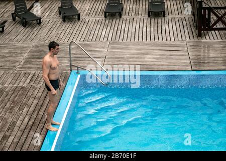 Junger Mann steht in der Nähe des blauen Beckens, bevor er ins Wasser springt. Nass weißer Kerl aus sportlichem Aufbau in Schwimmstämmen. Tourismus, Urlaubskonzept. Outd Stockfoto